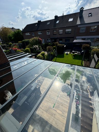 Gartenansicht mit Glasdach, Rasen, Hecken und Blumenbeeten. Heller, sonniger Himmel.